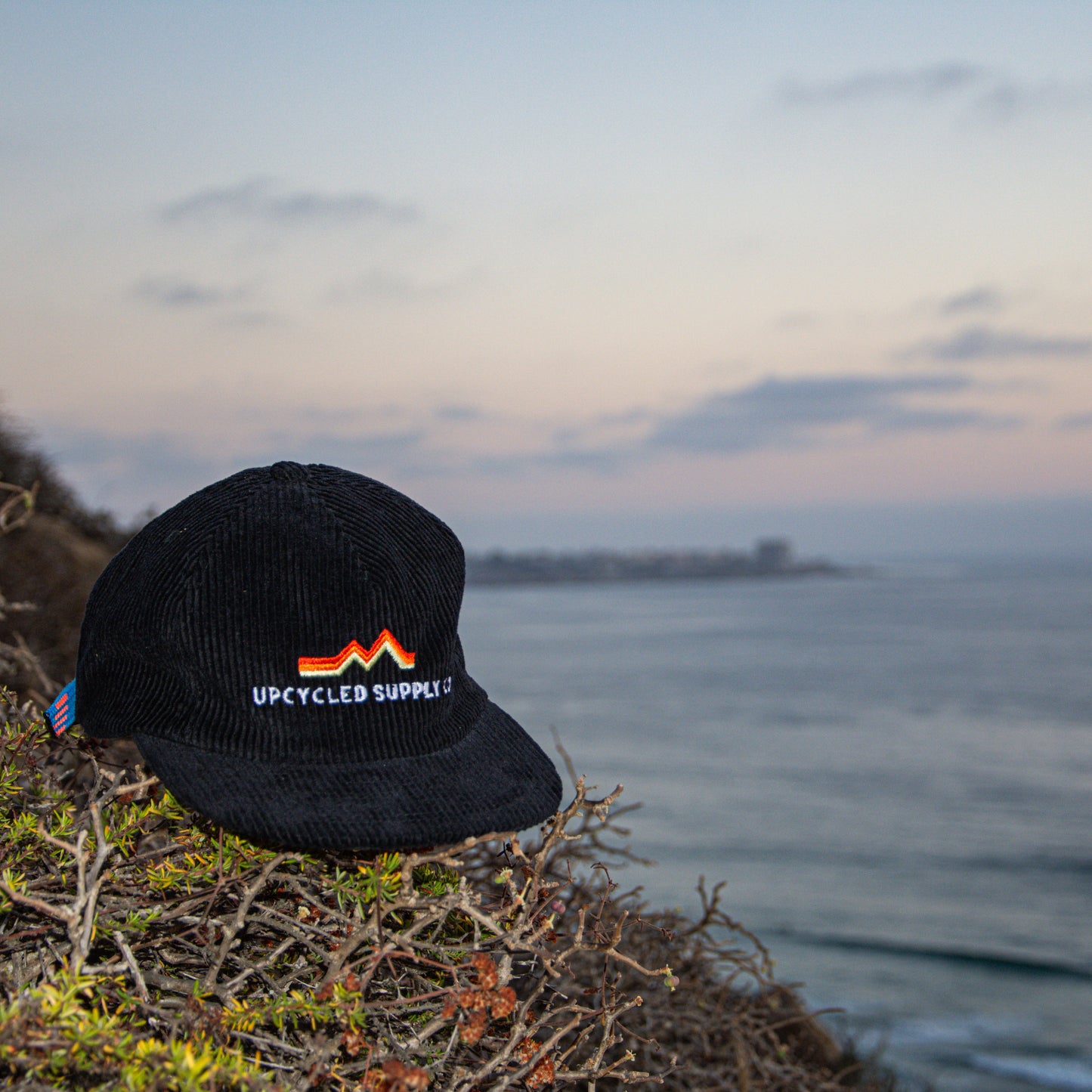 Black cap with 'Upcycled Supply' logo on a cliff overlooking the ocean.