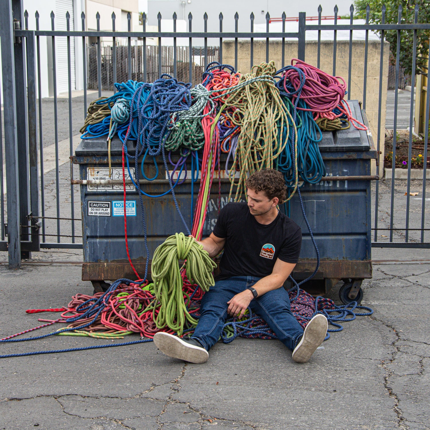 Man sitting on the ground next to a blue dumpster filled with colorful ropes.