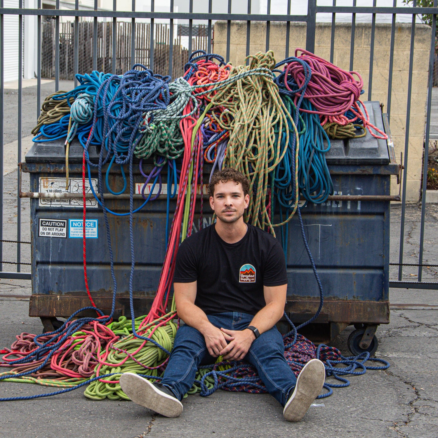 Man sitting in front of a dumpster filled with colorful ropes on a street.