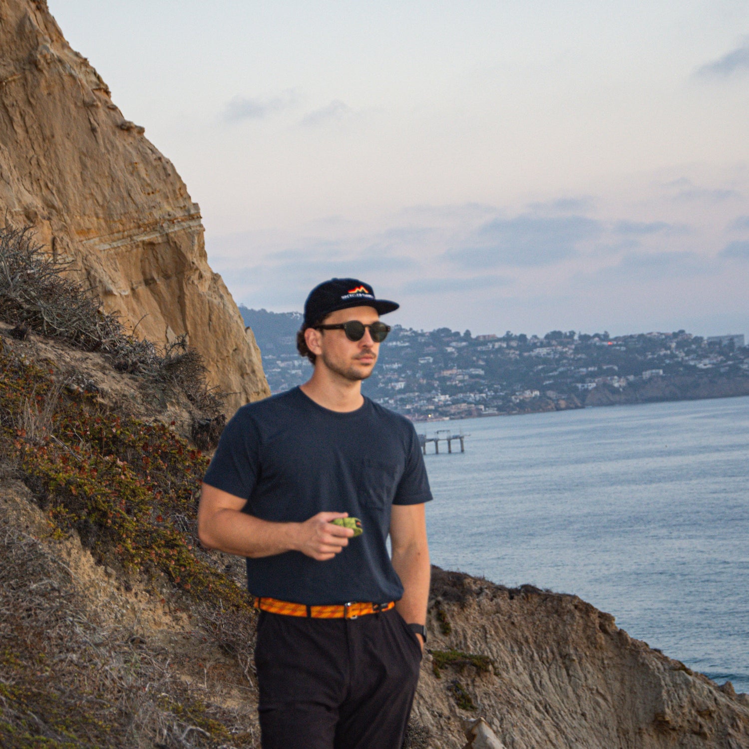 Man wearing an upcycled climbing rope belt and hat while standing on a coastal trail at sunset.