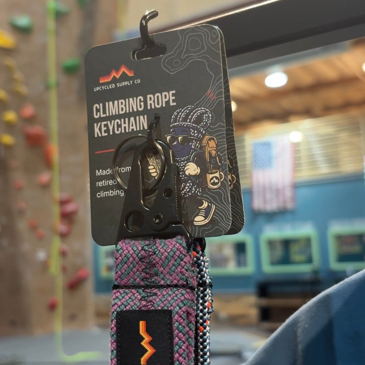 A recycled rope climbing rope keychain from Upcycle Supply Co. hanging from its black retail card tag, set against a blurred indoor climbing gym background.