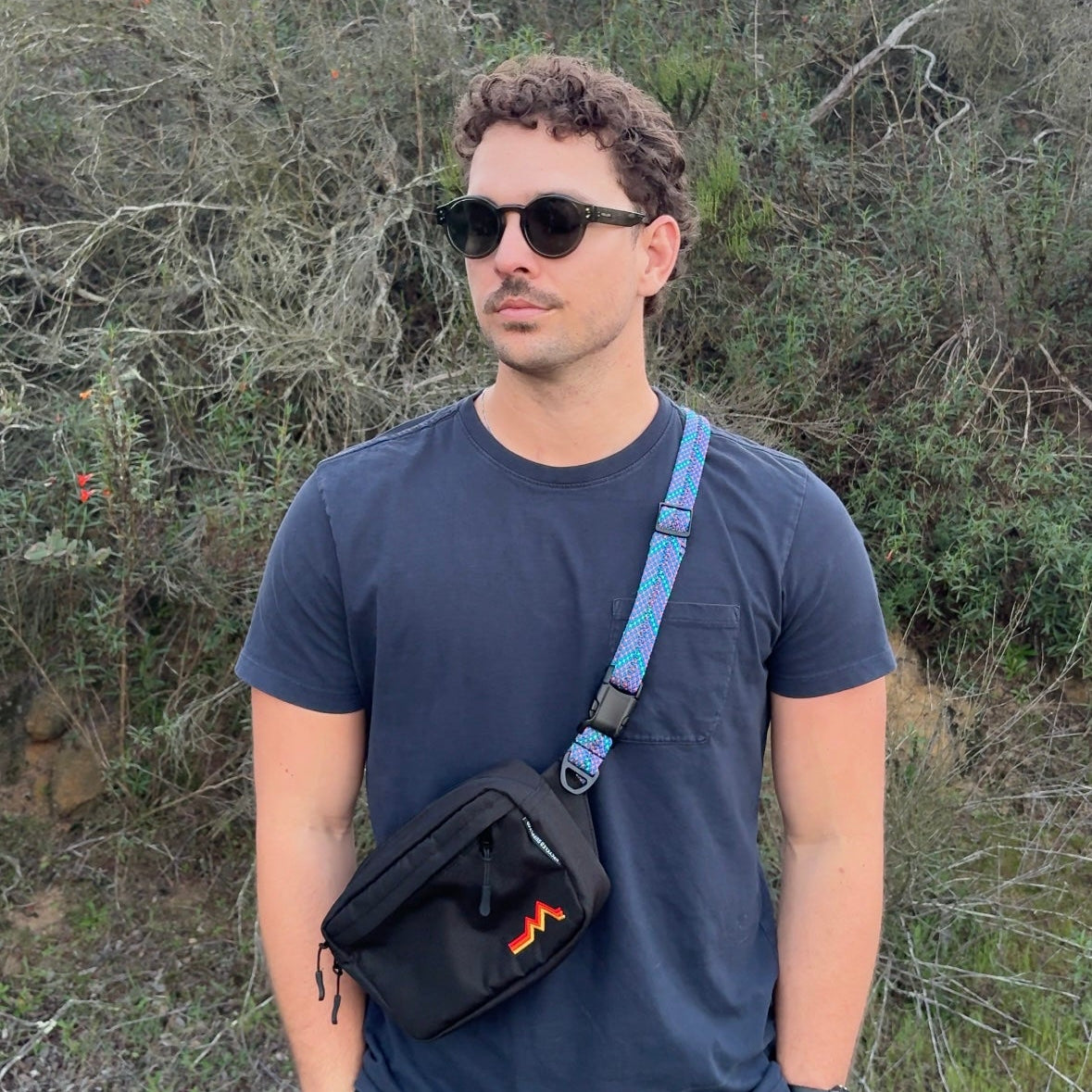 Man wearing sunglasses and a blue shirt with a black bag over his shoulder, standing in a natural setting.