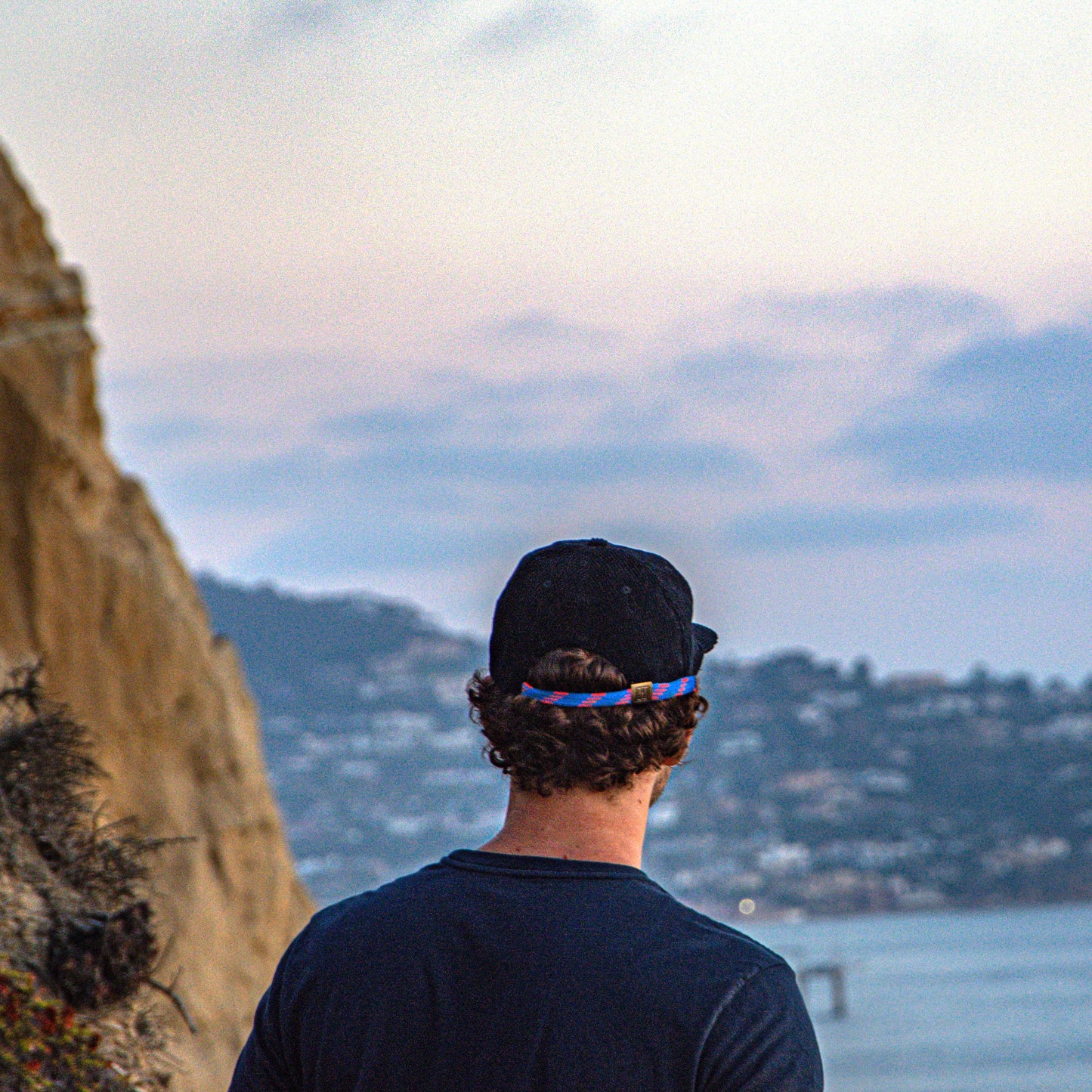 Person wearing a black cap and dark shirt looking at a scenic view of a coastal landscape with mountains and water.