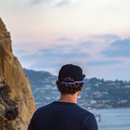 Person wearing a black cap and dark shirt looking at a scenic view of a coastal landscape with mountains and water.