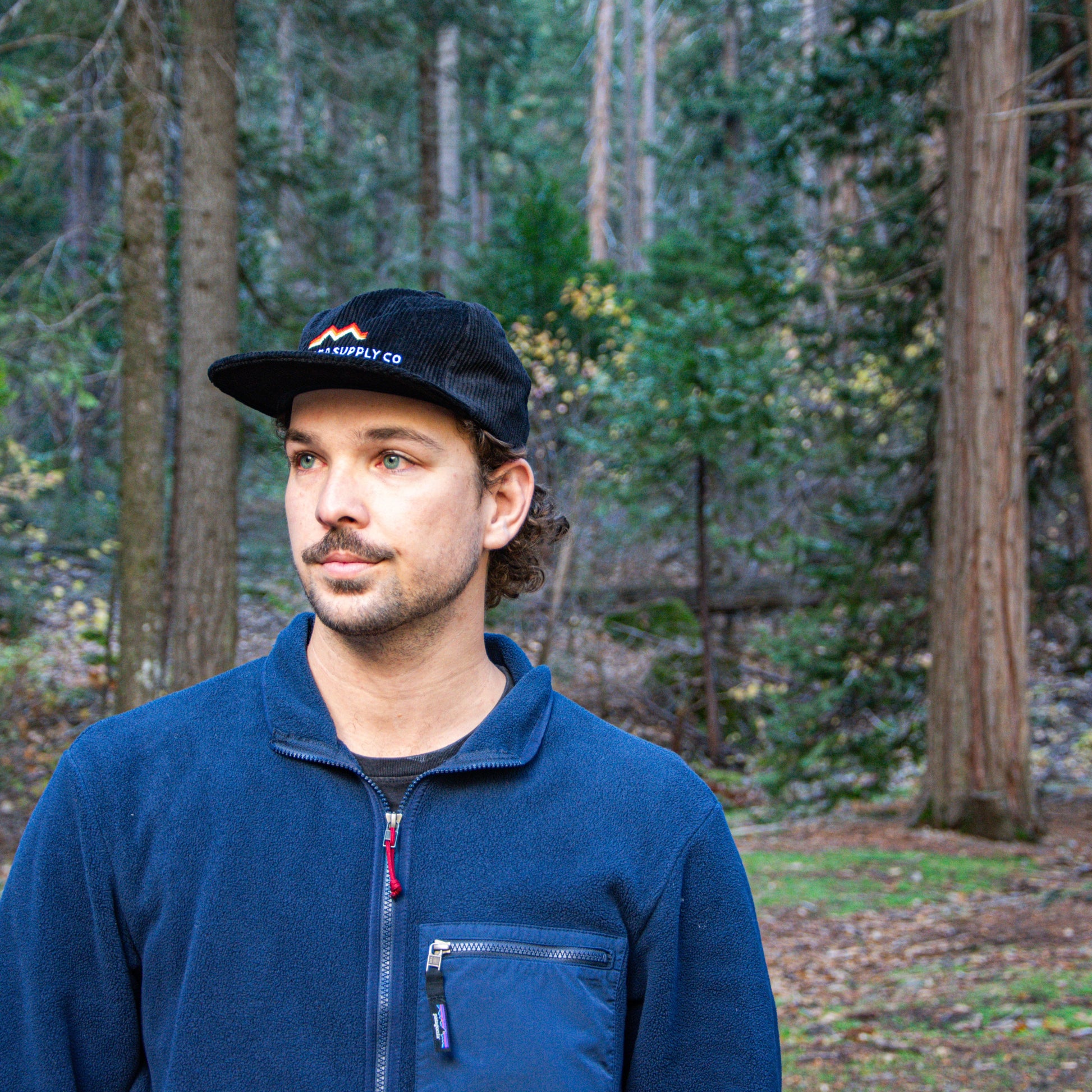 Man wearing a blue jacket and black cap with a logo, standing in a forest.