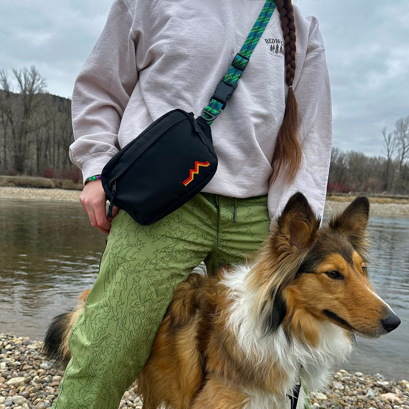 Person with a dog by a body of water, wearing a white sweatshirt and green pants.