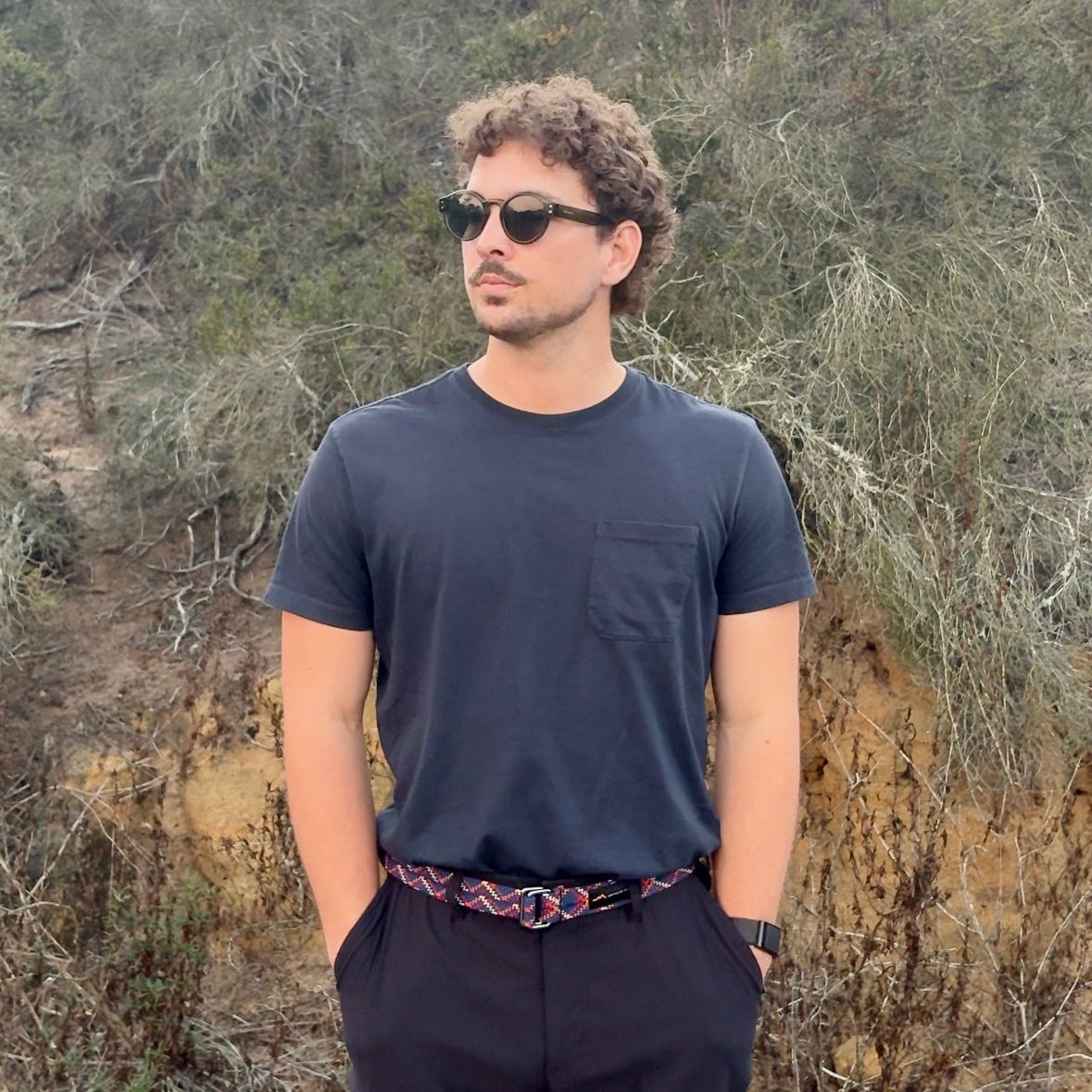 Man wearing sunglasses and a dark shirt standing in a natural setting with trees and shrubs.