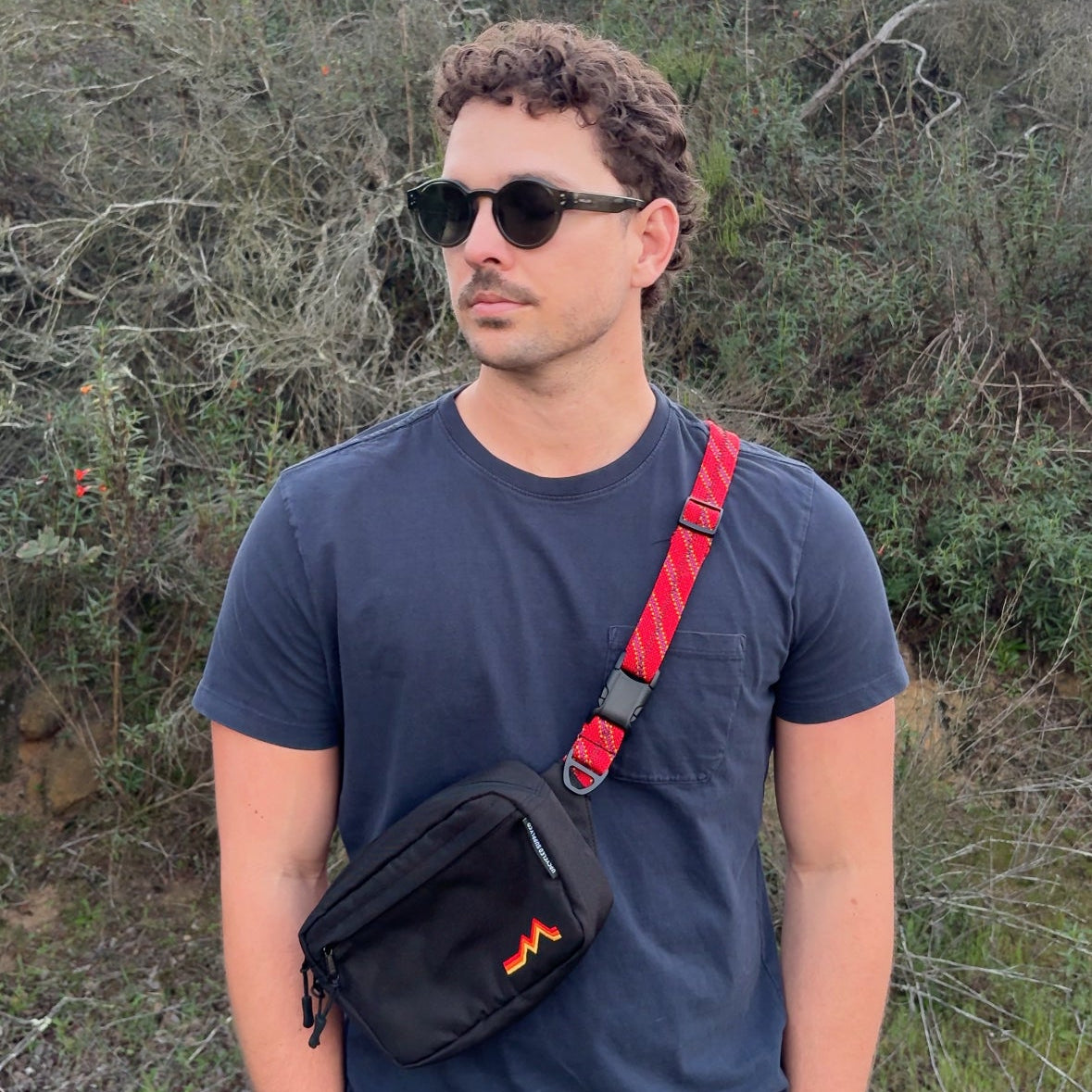 Man wearing sunglasses and a black shirt with a red strap, standing in a natural setting.