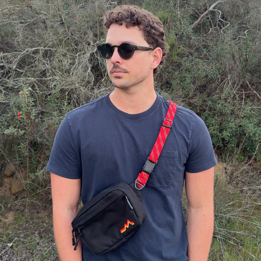 Man wearing sunglasses and a black shirt with a red strap, standing in a natural setting.