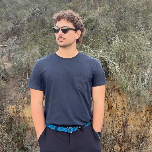 Man wearing sunglasses and a dark shirt standing in a natural setting with trees and shrubs.