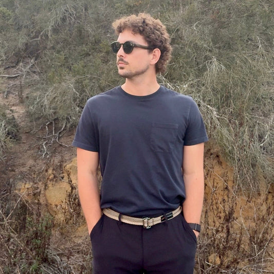 Man wearing sunglasses and a dark outfit standing in a natural setting with trees and shrubs.
