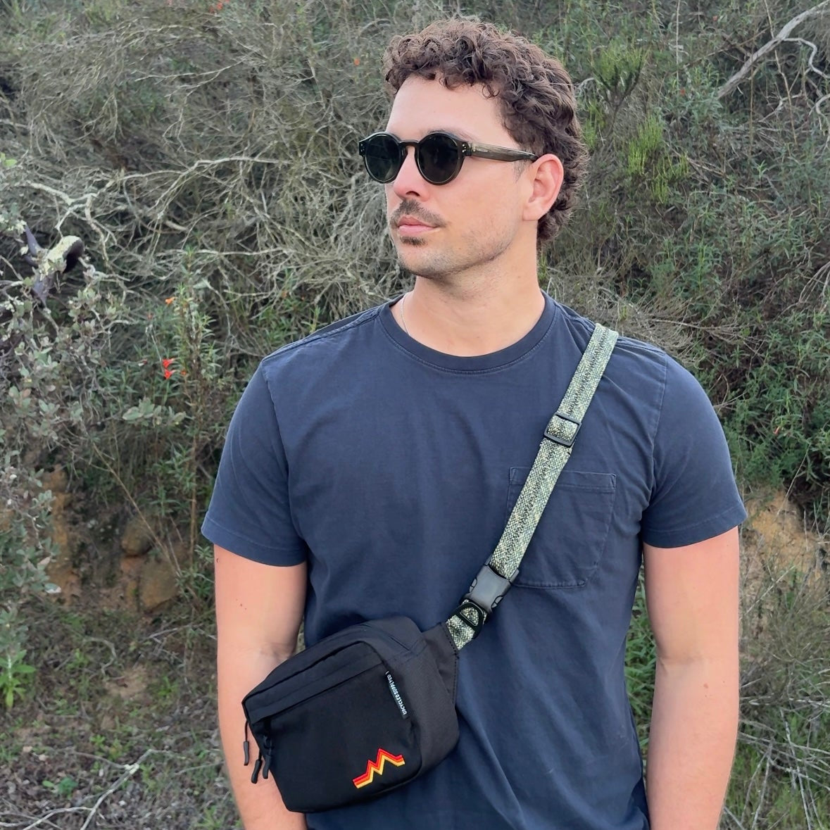 Man wearing sunglasses and a dark blue t-shirt with a crossbody bag in a natural setting