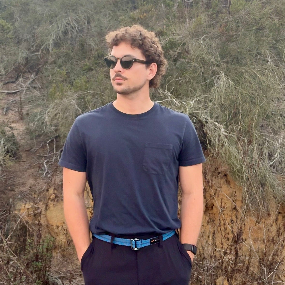 Man wearing sunglasses and a dark outfit standing in a natural setting with trees and shrubs.