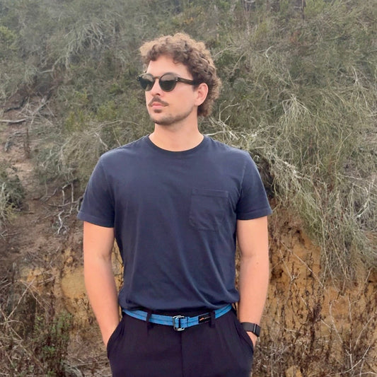 Man wearing sunglasses and a dark outfit standing in a natural setting with trees and shrubs.