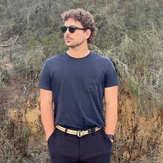 Man wearing sunglasses and a dark outfit standing in a natural setting with trees and shrubs.