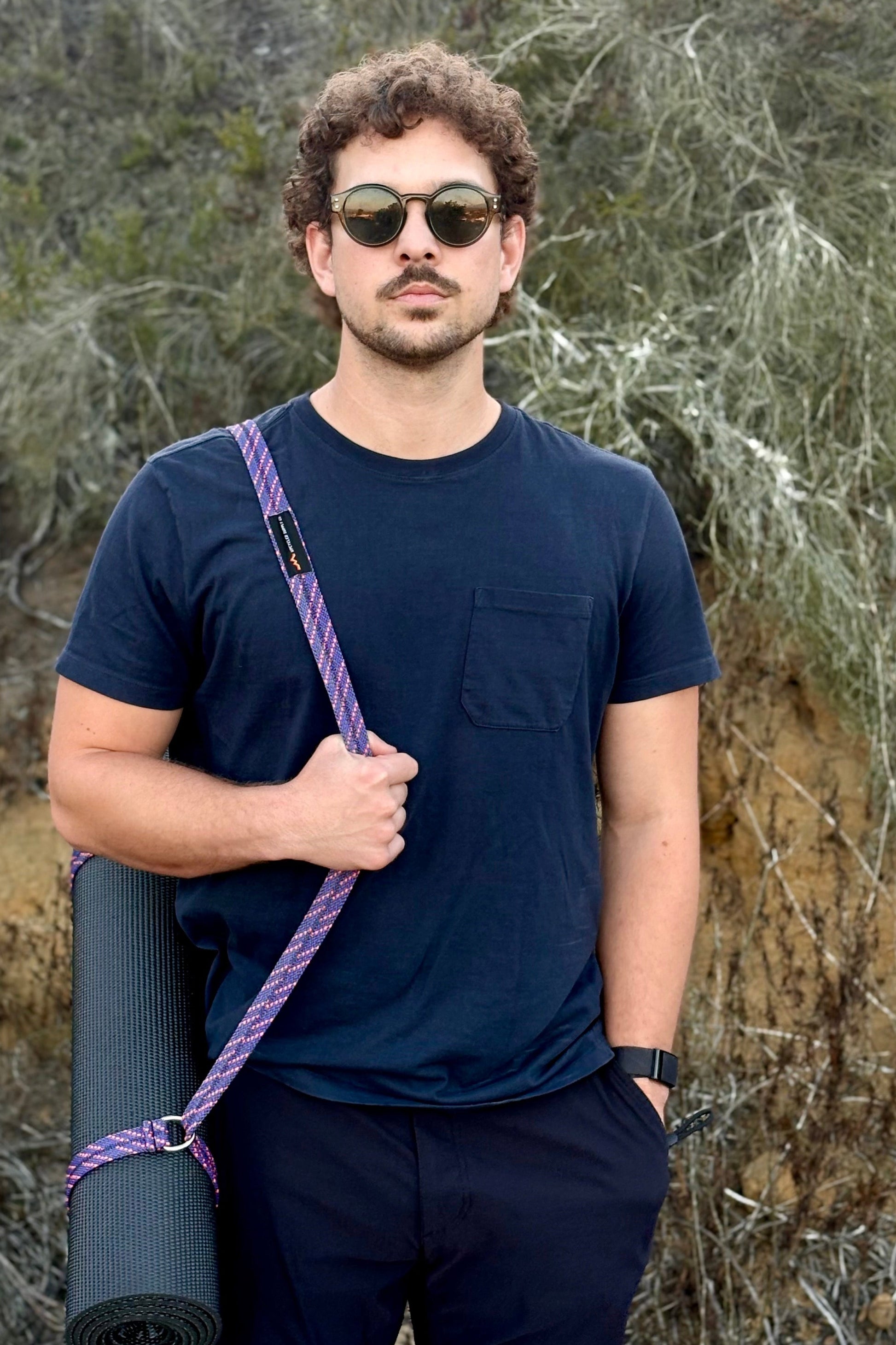Man holding a yoga mat with a purple strap against a natural background