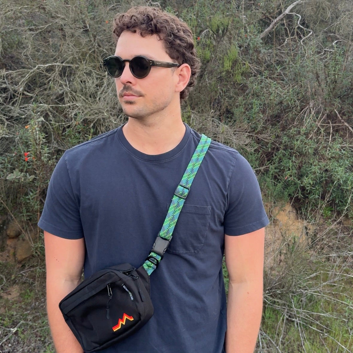 Man wearing sunglasses and a dark blue t-shirt with a green crossbody bag, standing in a natural setting.