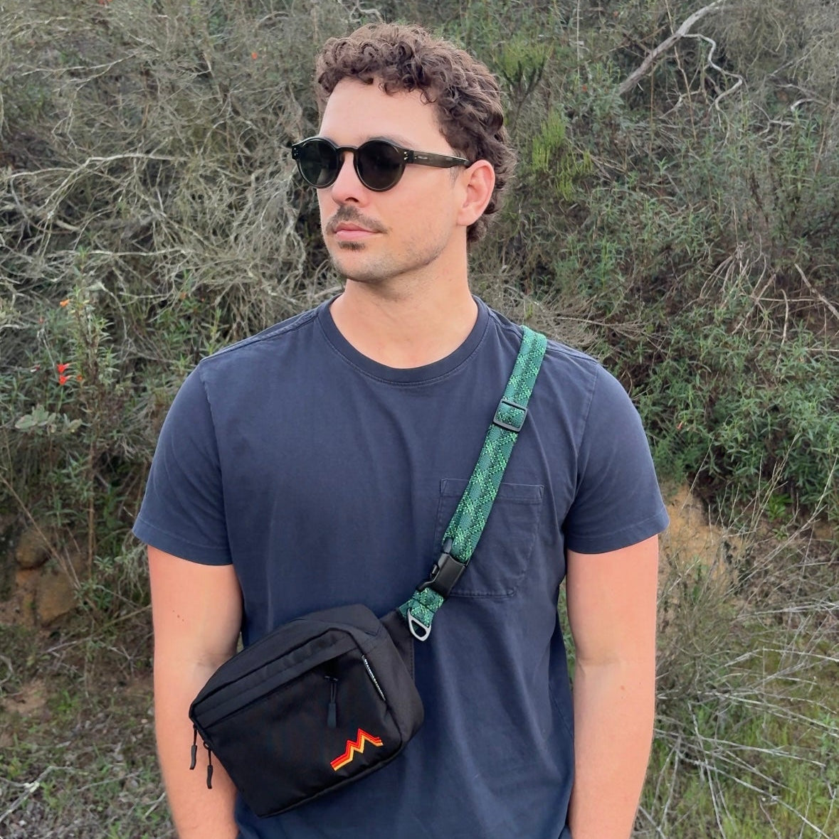 Man wearing sunglasses and a dark blue t-shirt with a green strap over his shoulder, standing in a natural setting.