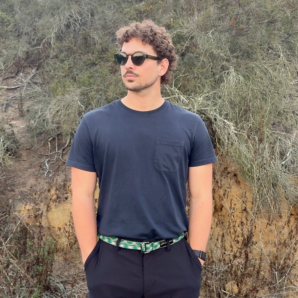 Man wearing sunglasses and a dark shirt standing in a natural setting with trees and rocks.