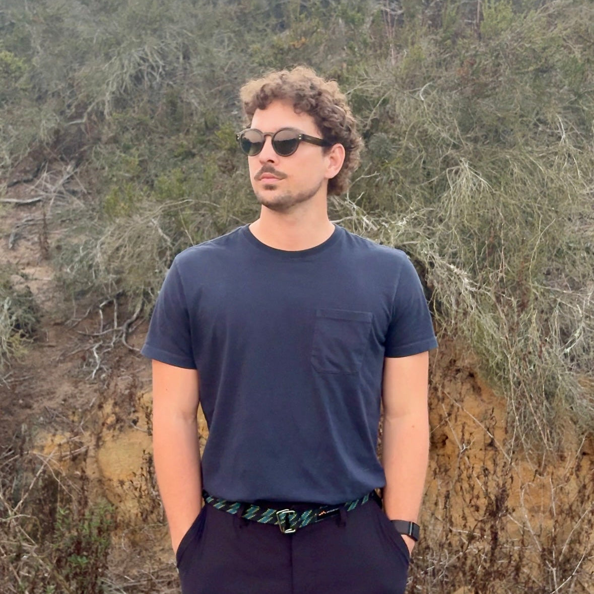 Man wearing sunglasses and a dark shirt standing in a natural setting with greenery.