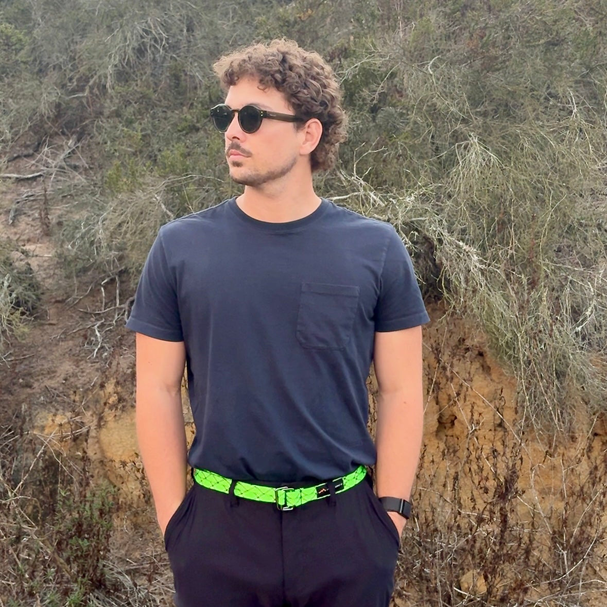 Man wearing sunglasses and a dark shirt with a green belt standing in a natural setting.