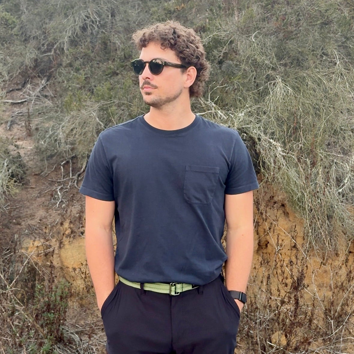 Man wearing sunglasses and a dark shirt standing in a natural setting with trees and rocks.