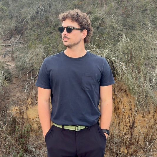 Man wearing sunglasses and a dark shirt standing in a natural setting with trees and rocks.