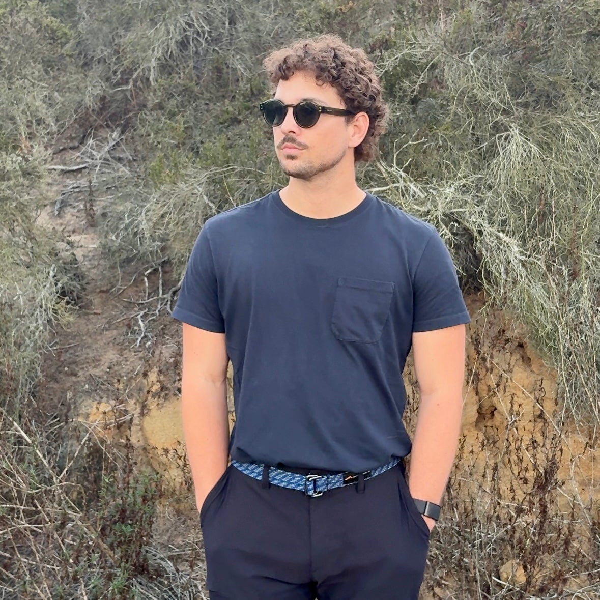Man wearing sunglasses and a dark outfit standing in a natural setting with trees and shrubs.
