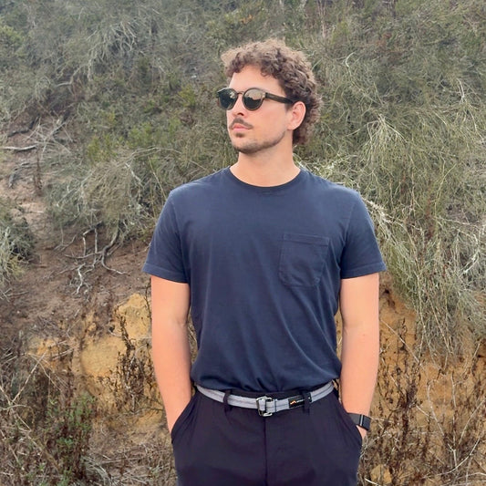 Man wearing sunglasses and a dark shirt standing in a natural setting with trees and rocks.