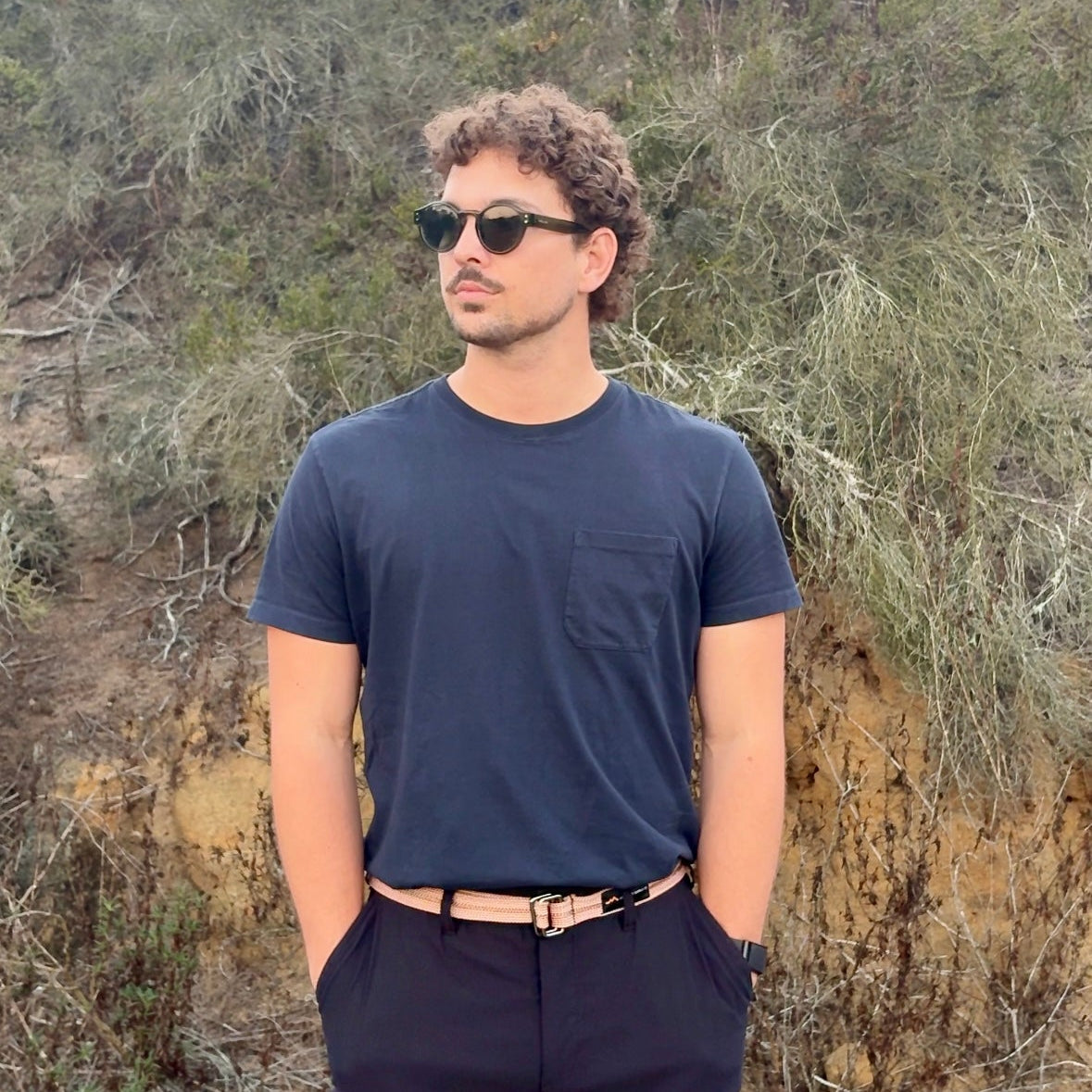 Man wearing sunglasses and a dark outfit standing in a natural setting with trees and rocks.